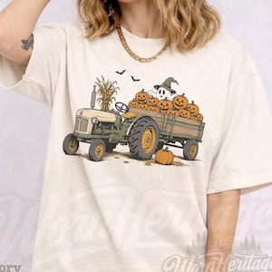 May include: A cream-colored t-shirt featuring a graphic of a green tractor pulling a wooden trailer filled with orange jack-o'-lanterns and a white ghost wearing a green hat. Two small black bats fly above the trailer, and a single pumpkin sits on the ground.