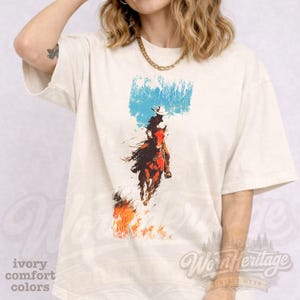 May include: Cream-coloured t-shirt with a vintage cowboy and horse graphic, set against a blue sky and flames. The shirt also features the text "ivory comfort colors". The brand name "Worn Heritage" is visible.