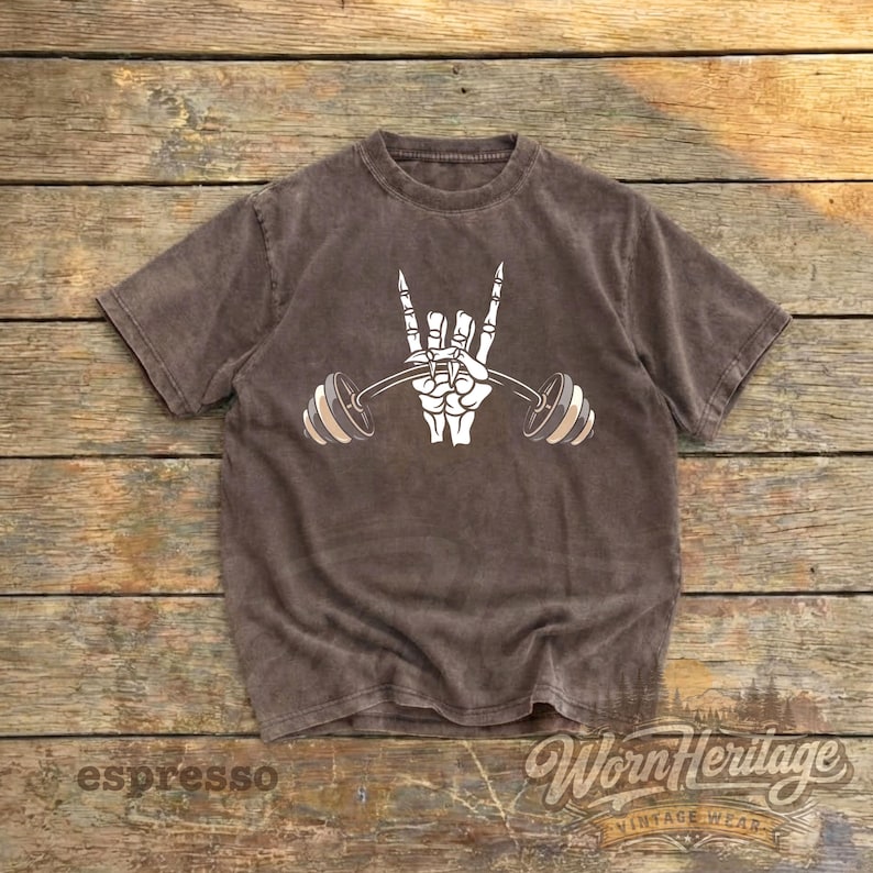 Peut inclure: T-shirt couleur expresso avec un motif blanc d'une main de squelette tenant une barre de musculation en signe de rock and roll. Le t-shirt a un aspect vintage et les mots "Worn Heritage" sont imprim&eacute;s en bas &agrave; droite.