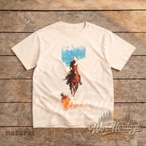 May include: Cream-coloured t-shirt with a graphic of a cowboy on horseback, set against a blue and white backdrop. The cowboy is riding through flames. The shirt features the text "Worn Heritage Vintage Wear" and "natural".