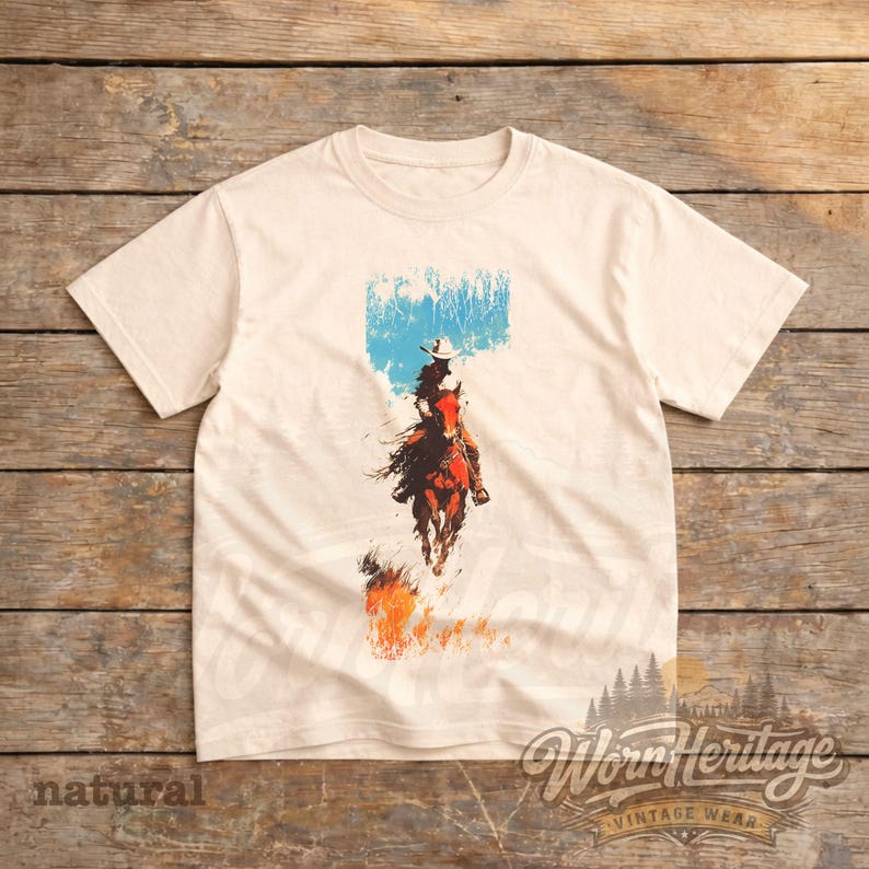 May include: Cream-coloured t-shirt with a graphic of a cowboy on horseback, set against a blue and white backdrop. The cowboy is riding through flames. The shirt features the text "Worn Heritage Vintage Wear" and "natural".