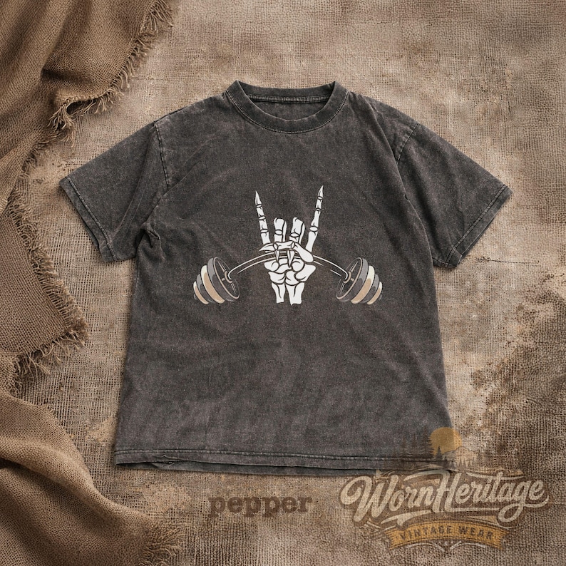 Peut inclure: T-shirt gris fonc&eacute; avec une finition vieillie. Le motif repr&eacute;sente une main de squelette blanche faisant le signe rock, tenant une barre. Le texte "pepper" est en bas &agrave; gauche et "Worn Heritage Vintage Wear" en bas &agrave; droite.