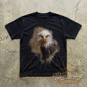 May include: Black t-shirt featuring a detailed graphic of a bald eagle's head and chest. The eagle is depicted with white feathers, a yellow beak, and a stern expression. The shirt has a vintage wear logo at the bottom.