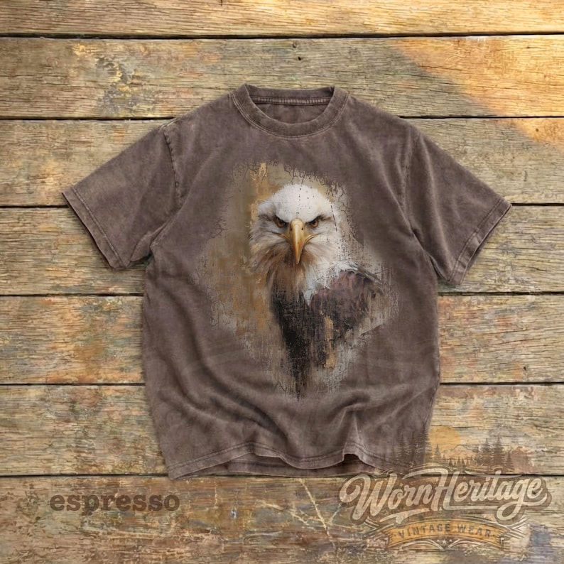 May include: Espresso-colored t-shirt featuring a detailed graphic of a bald eagle. The eagle's head is in sharp focus, with a white head, yellow beak, and intense gaze. The shirt has a vintage look, with the words "Worn Heritage" and "espresso" visible.