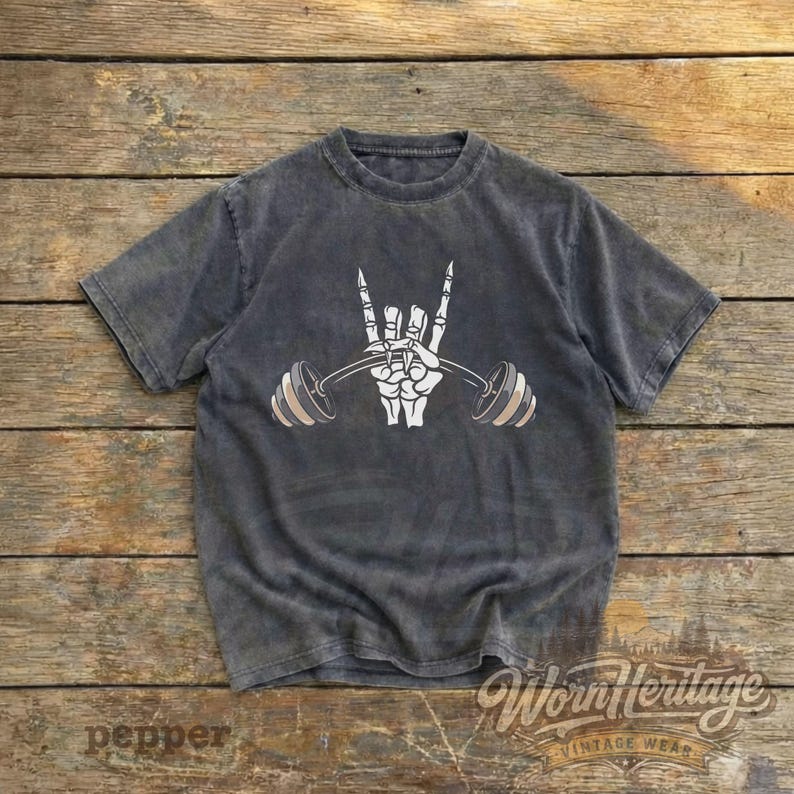 Peut inclure: T-shirt gris fonc&eacute; avec un motif de main de squelette blanche tenant une barre de musculation. Le design inclut le geste de la main rock and roll. Le t-shirt a un aspect vintage et us&eacute;. Le texte "Worn Heritage Vintage Wear" est en bas.
