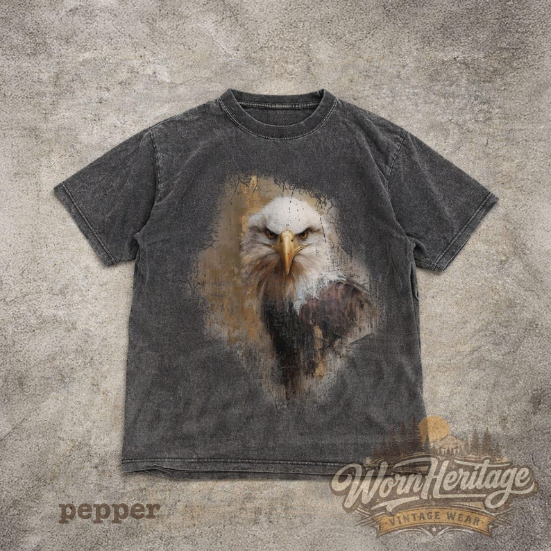 May include: A dark gray t-shirt featuring a detailed graphic of an eagle's face. The eagle is depicted with white, brown, and black feathers, and a yellow beak. The shirt has the text "pepper" at the bottom left and the "Worn Heritage Vintage Wear" logo.