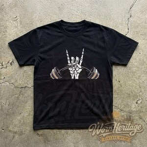 Peut inclure: T-shirt noir avec un motif de main de squelette blanche faisant le signe du rock and roll et tenant une barre de musculation. La barre a des poids beiges et gris. Le t-shirt a un col rond et des manches courtes.