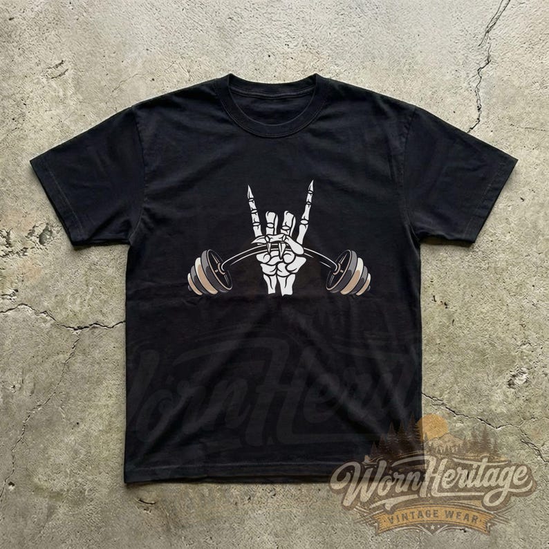 Peut inclure: T-shirt noir avec un motif de main de squelette blanche faisant le signe du rock and roll et tenant une barre de musculation. La barre a des poids beiges et gris. Le t-shirt a un col rond et des manches courtes.