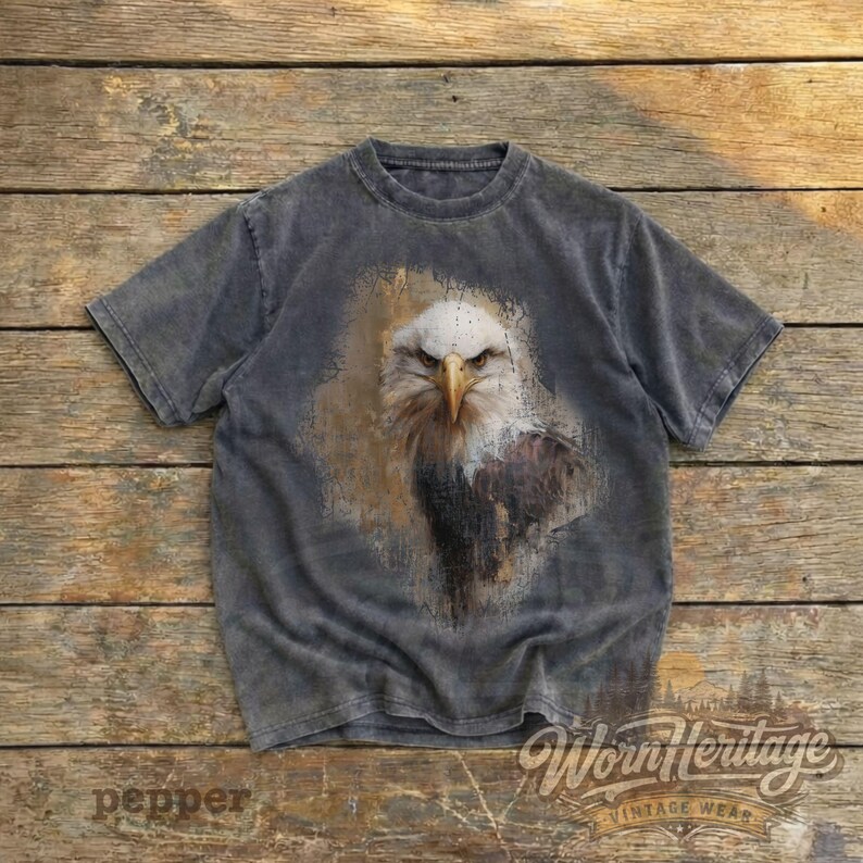 May include: A gray, short-sleeved t-shirt featuring a detailed graphic of an eagle's head. The eagle is depicted with white, brown, and gray tones, set against a textured background. The shirt has the text "Worn Heritage Vintage Wear".