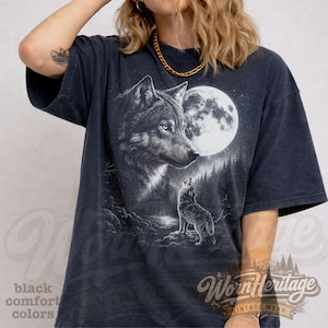 Op de afbeelding: Donkergrijze t-shirt met een afbeelding van een wolf die naar de maan huilt. Het ontwerp bevat een volle maan, bomen en een tweede wolf. Het shirt heeft de tekst "Worn Heritage" en "black comfort colors".