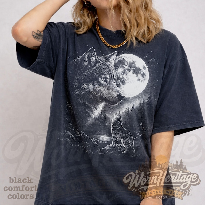 Op de afbeelding: Donkergrijze t-shirt met een afbeelding van een wolf die naar de maan huilt. Het ontwerp bevat een volle maan, bomen en een tweede wolf. Het shirt heeft de tekst "Worn Heritage" en "black comfort colors".