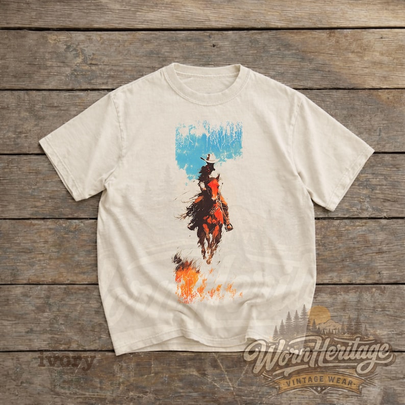 May include: Ivory t-shirt with a vintage graphic of a cowboy on horseback. The design includes a blue sky, flames, and the words "Worn Heritage Vintage Wear". The shirt is a classic crew neck.