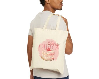 Puede incluir: Bolso tote de color crema con un gráfico rosa de una tarta en capas con una cereza y las palabras "Cherry on Top". La bolsa tiene forma rectangular y dos correas largas.
