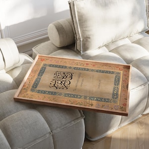May include: A rectangular wooden tray with ornate calligraphy and floral border designs. The tray features a beige background with black text and is resting on a light gray sofa. The text reads "So surely with hardship comes ease."