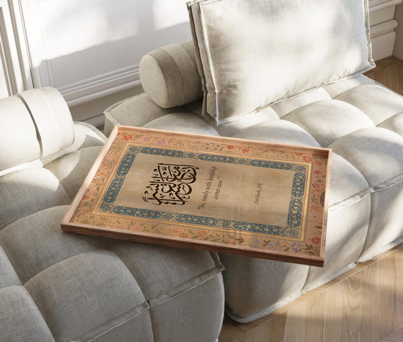 May include: A rectangular wooden tray with ornate calligraphy and floral border designs. The tray features a beige background with black text and is resting on a light gray sofa. The text reads "So surely with hardship comes ease."