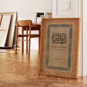 May include: A framed print with Arabic calligraphy and English text, "So, surely with hardship comes ease." The frame is wood-toned with floral and geometric border designs. The print is displayed on a wooden floor.