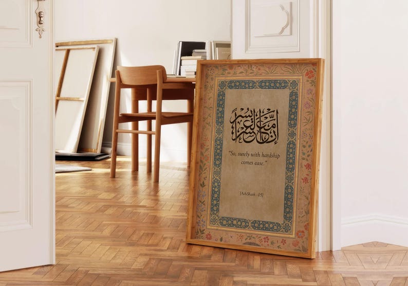 May include: A framed print with Arabic calligraphy and English text, "So, surely with hardship comes ease." The frame is wood-toned with floral and geometric border designs. The print is displayed on a wooden floor.