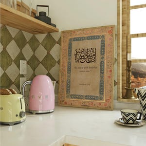 May include: A kitchen scene featuring a pink electric kettle and a yellow toaster. A framed artwork with Arabic calligraphy and the text "So, surely with hardship comes ease" is displayed. A teacup and a decorative vase are also visible.
