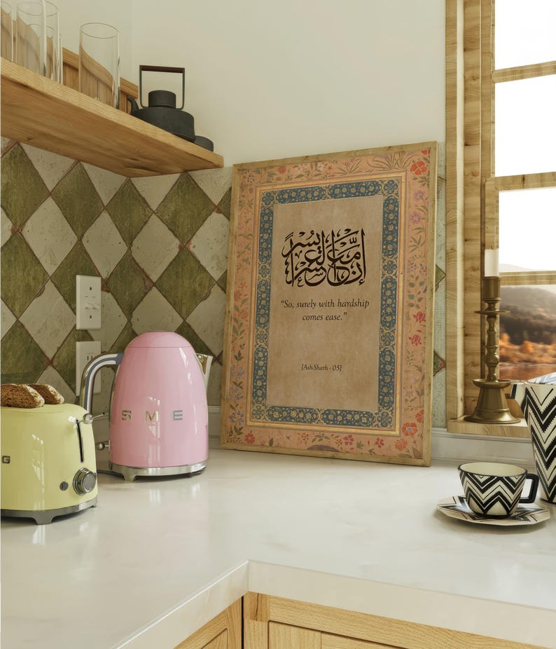 May include: A kitchen scene featuring a pink electric kettle and a yellow toaster. A framed artwork with Arabic calligraphy and the text "So, surely with hardship comes ease" is displayed. A teacup and a decorative vase are also visible.