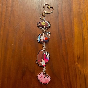 May include: A gold-colored keychain featuring four cartoon character charms. The charms include a brown box with a gold "M", a boy hanging upside down, a girl hanging upside down, and a pink pig. The keychain has a heart-shaped clasp.