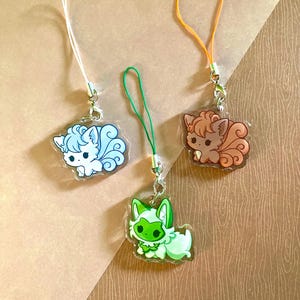 May include: Three acrylic charms featuring cartoon foxes in blue, brown, and green. Each charm has a different colored strap and a silver-colored clasp. The foxes have fluffy tails and are in a cute, cartoon style.