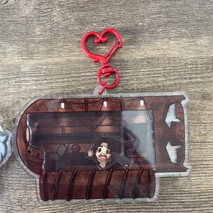 Puede incluir: Un llavero acrílico transparente con una ilustración de dibujos animados de una persona dentro de un marco marrón de temática industrial. El llavero tiene un cierre rojo en forma de corazón. La ilustración muestra a una persona saludando con una expresión de sorpresa.