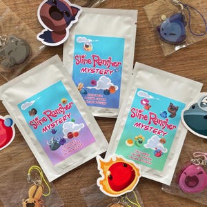 Puede incluir: Se muestran varios paquetes blancos con el texto "Slime Rancher MYSTERY", cada uno con un diseño colorido con slimes y nubes de dibujos animados. Los paquetes incluyen un dije y una pegatina de slime. Varios dijes y pegatinas con temática de slime están dispersos.
