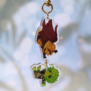 Puede incluir: Un dije de acrílico de dos niveles con dos personajes ilustrados que cuelgan de un broche dorado en forma de corazón. El dije superior muestra una figura con armadura dorada y capa roja oscura, mientras que el inferior muestra un personaje verde.