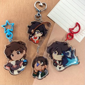 Puede incluir: Cuatro llaveros de acrílico transparente con ilustraciones de dibujos animados de un personaje con diferentes expresiones y poses. El personaje lleva diferentes atuendos en cada llavero. Los llaveros están unidos a anillos y clips metálicos.