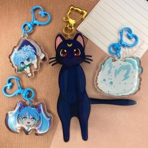 Pode incluir: Quatro chaveiros diferentes com personagens de anime. Os chaveiros são feitos de acrílico transparente e apresentam um gato azul, uma menina de cabelo azul, um menino de cabelo azul e uma criatura branca e fofa. Os chaveiros são presos a anéis azuis em forma de coração.