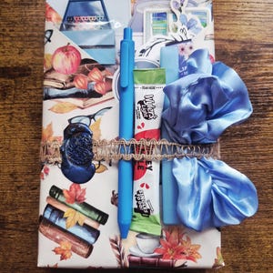 May include: A rectangular package wrapped in paper featuring illustrations of books, apples, and autumn leaves. It is secured with a burlap ribbon, a blue wax seal, a blue pen, a Wyler's Light drink mix packet, and a light blue satin hair scrunchie.