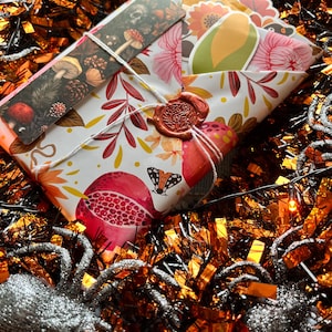 Puede incluir: Un regalo envuelto con un patrón floral y de granada, sellado con un sello de cera y atado con hilo. Un marcador con ilustraciones de hongos está en la parte superior. El regalo está rodeado de oropel naranja y negro y una araña brillante.