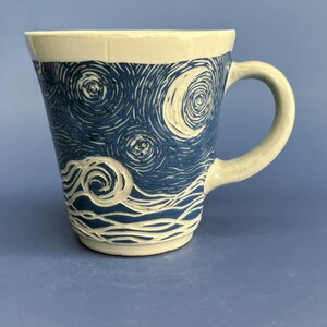 May include: A ceramic mug with a cream-colored base and a deep blue hand-carved design featuring swirling patterns, circular stars, and a crescent moon. The mug has a wide rim, a tapered body, and a curved handle on the right side of the vessel.