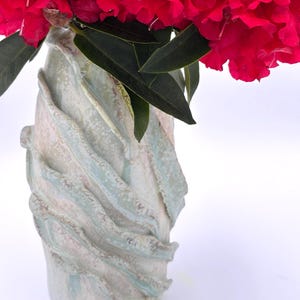 Puede incluir: Un jarrón de cerámica con un diseño texturizado y arremolinado en tonos blancos, azules y grises sostiene un vibrante ramo de flores rojas brillantes y hojas verdes. El jarrón mide unos 25 cm de alto.