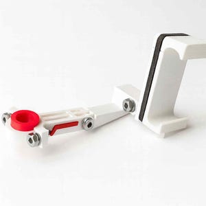May include: A white plastic device with a red circular component and a black padded grip. The device is assembled with silver bolts and has a rectangular base. The design appears to be for holding an object.