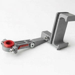 Może przedstawiać: Szare, regulowane urządzenie z czerwonym okrągłym elementem i srebrnymi śrubami. Urządzenie posiada czarny pasek i prostokątny uchwyt. Urządzenie jest przeznaczone do trzymania przedmiotu.