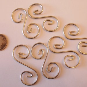 May include: A set of eight silver metal ornament hooks with a decorative swirl design. The hooks are arranged in a pattern on a white surface.