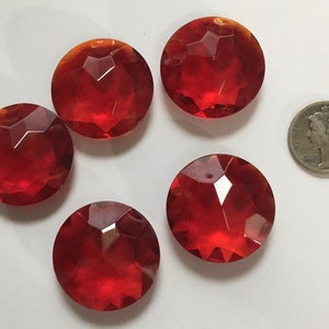 May include: Five faceted, round, red glass gems. The gems are arranged on a white surface, with a silver coin for size comparison. The gems have a brilliant, transparent appearance, with multiple facets catching the light.