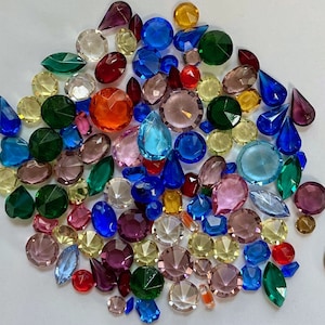 May include: A collection of colorful glass gems in various shapes and sizes, including round, square, and star-shaped gems. The gems are arranged in a random pattern on a white surface.