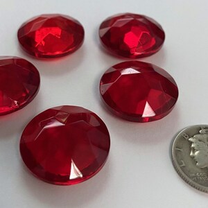 May include: Six faceted, oval-shaped, bright red gemstones are displayed, alongside a silver coin. The gems have a brilliant, reflective surface. The coin is a Mercury dime, dated 1941.