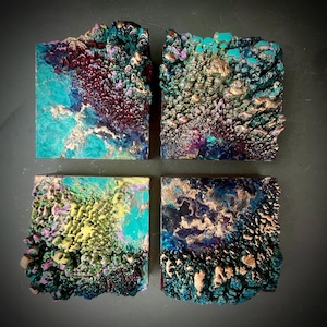 May include: Four square abstract art pieces with textured surfaces. Each piece features a unique combination of colors, including teal, gold, purple, and yellow. The art has a three-dimensional appearance, with raised areas and intricate details.