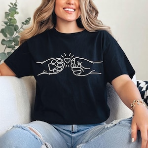 Op de afbeelding: Een zwart t-shirt met korte mouwen en een witte lijntekening op de borst. Het ontwerp toont een menselijke vuist die een boks geeft tegen een dierenpoot, met een klein hartje direct boven het contactpunt tussen de twee figuren.