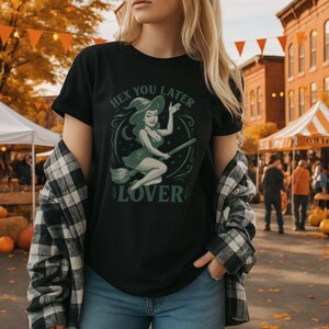 May include: Black t-shirt featuring a green and white illustration of a witch on a broom, with the text "HEX YOU LATER LOVER". The witch is wearing a hat and dress. The shirt is worn with a plaid shirt and blue jeans.