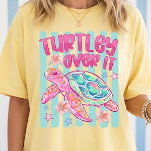 May include: Yellow short-sleeved t-shirt with the text "TURTLEY OVER IT" above a colorful turtle graphic. The turtle has a pink body, a teal shell, and is surrounded by flowers and stars. The shirt is a casual style.
