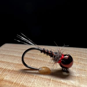 May include: Close-up of a fishing fly with a red metallic bead head, a dark brown body, and a curved hook. The fly has a small, tan-colored bead at the hook's bend and several thin, silver-colored fibers extending from the top.