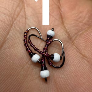 May include: A handcrafted, decorative earring featuring a dark purple wire wrapped around a silver metal frame. The earring has three white and black beads. The earring is a unique piece of jewelry.