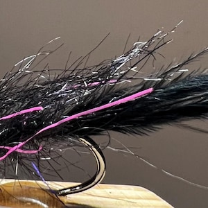 May include: A black and silver fishing fly with a silver bead head and a curved hook. The fly has black feathers and fibers with pink and silver accents. The fly is designed for fishing.
