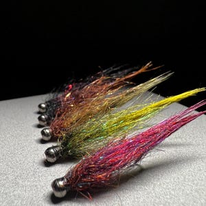 May include: Five colorful fishing flies with silver ball heads. The flies are arranged in a row, showcasing a gradient of colors including red, yellow, green, and brown. The flies are designed with iridescent materials.