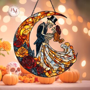 Pode incluir: Suncatcher de vitral com um casal de esqueletos abra&ccedil;ados dentro de uma lua crescente. O design incorpora rosas vermelhas e amarelas, com uma corrente preta para pendurar. A imagem tem um tema do Dia dos Mortos, com ab&oacute;boras e uma caveira.