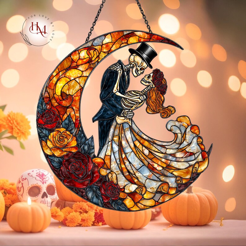 Pode incluir: Suncatcher de vitral com um casal de esqueletos abra&ccedil;ados dentro de uma lua crescente. O design incorpora rosas vermelhas e amarelas, com uma corrente preta para pendurar. A imagem tem um tema do Dia dos Mortos, com ab&oacute;boras e uma caveira.